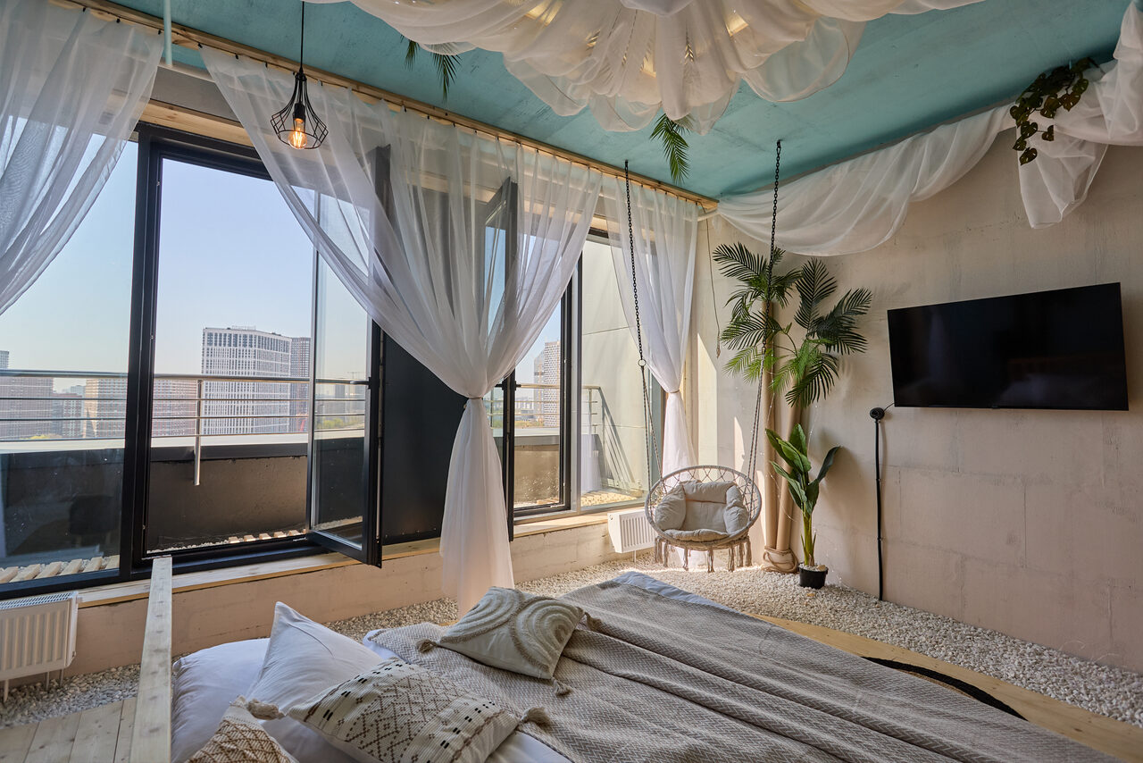 Арендовать Лофт Мальдивы с открытым душем, балконом и видом на Сити. Спальня с кроватью, окнами в пол и открытой дверью на балкон. Воздушные белые шторы, декоративные растения, светлая постель. Светло-голубой потолок с текстильными элементами. Телевизор на правой стене, подвесная лампа. Бежевые стены и пол с ковром.