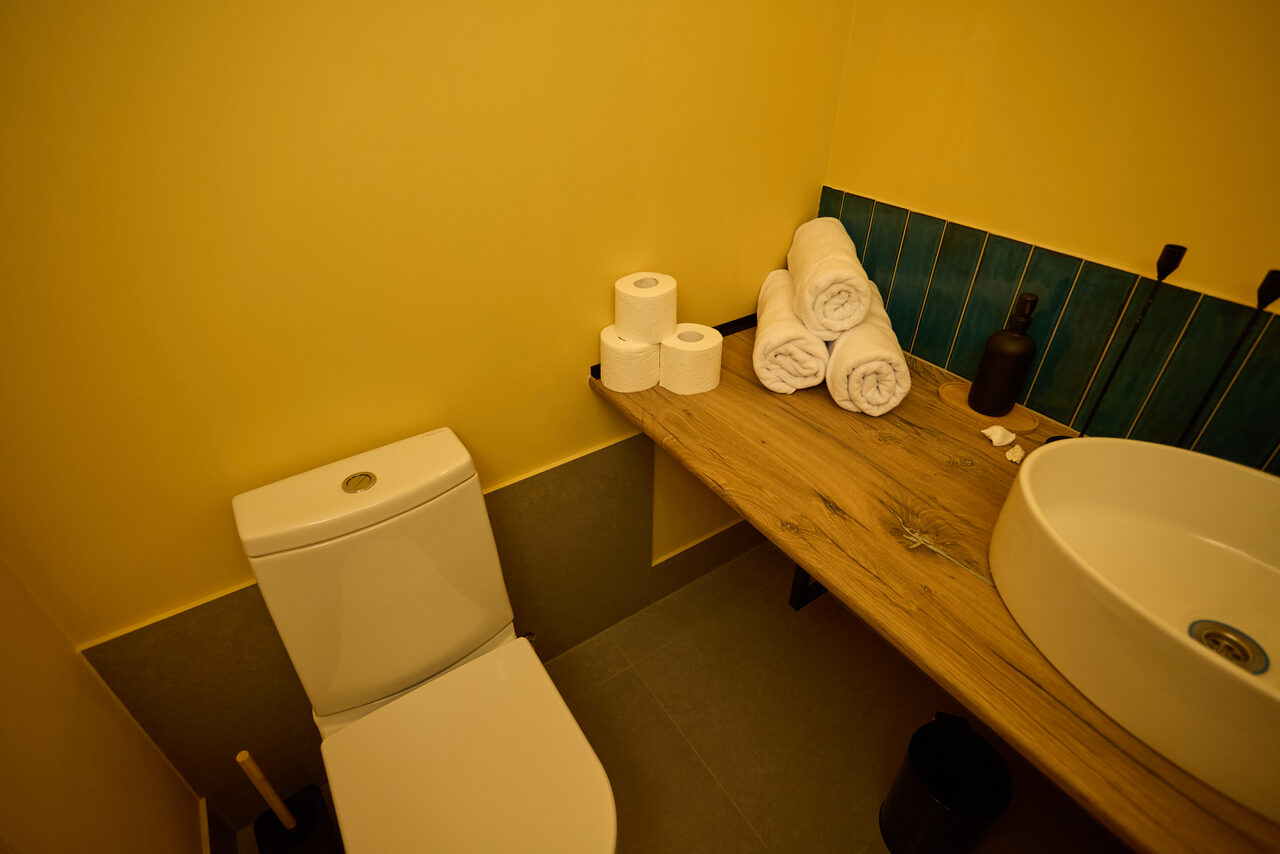 Арендовать Лофт Мальдивы с открытым душем, балконом и видом на Сити. Туалетная комната с жёлтыми стенами и серым полом, унитаз, деревянная столешница с раковиной круглой формы, три рулона туалетной бумаги, сложенные полотенца, чёрный дозатор, синие керамические плитки на стене, мусорное ведро.