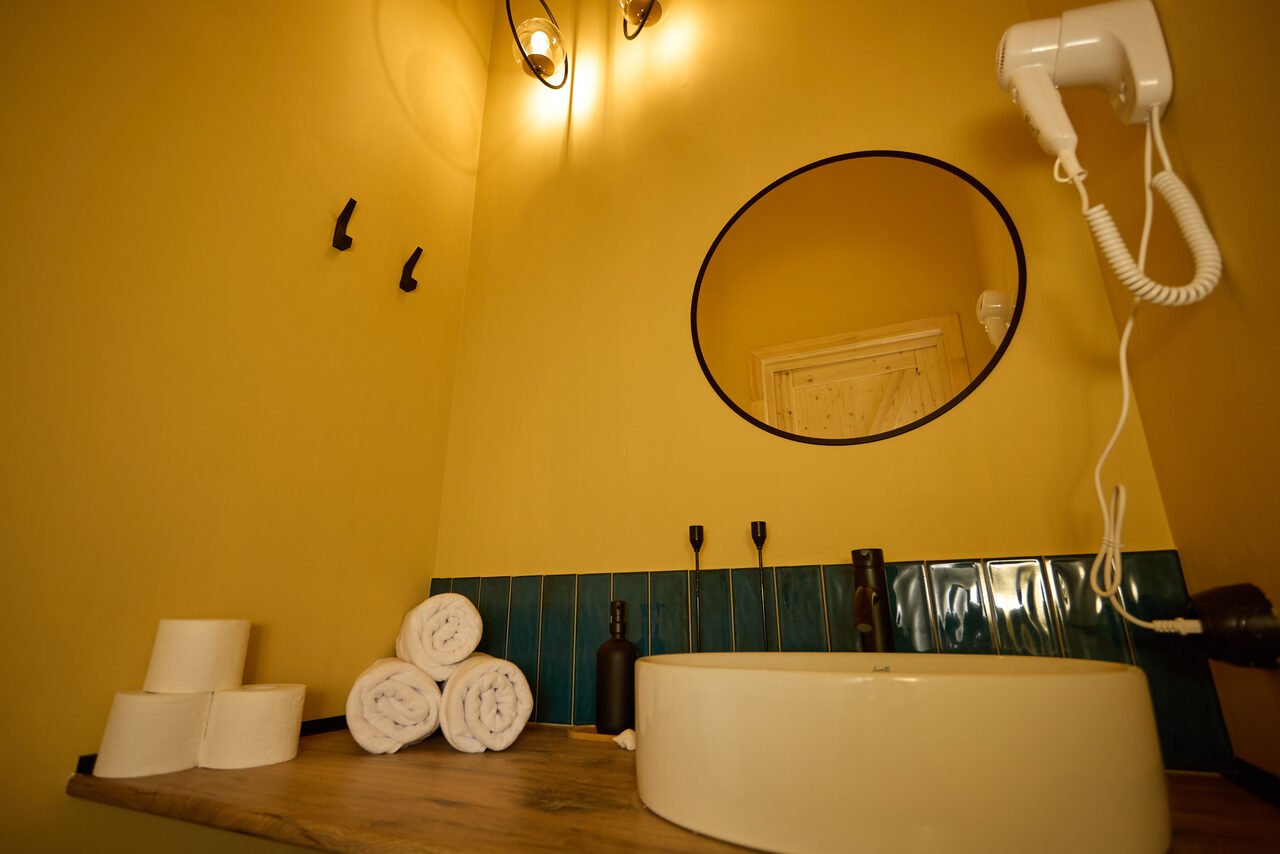 Арендовать Лофт Мальдивы с открытым душем, балконом и видом на Сити. Ванная комната с круглым зеркалом над раковиной, желтые стены, синие плитки на уровне раковины, деревянная столешница, фен, три крючка на стене, сложенные белые полотенца, несколько рулонов туалетной бумаги, светильники на стене.