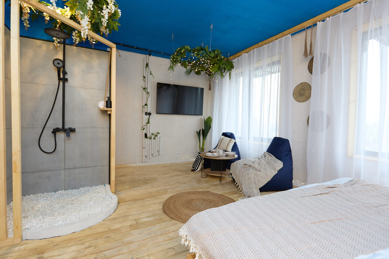 Арендовать Лофт Бали с фото зоной на 23 этаже с видом на Москва-реку. Комната с синем потолком и деревянным полом. Открытая душевая с черной сантехникой. Белые шторы на окнах, кресло-мешок, небольшой стол, подвешенные растения. На стене телевизор, декор в виде зелени и тарелок. Светлое покрывало на кровати.