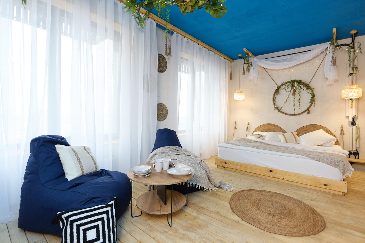 Арендовать Лофт Бали с фото зоной на 23 этаже с видом на Москва-реку. Спальня с двухместной кроватью, двумя подвесными светильниками, круглым деревянным столом и синим креслом-мешком. Белые стены и занавески, синий потолок, деревянный пол. Декор из текстиля и растений, круглое зеркало в изголовье кровати.