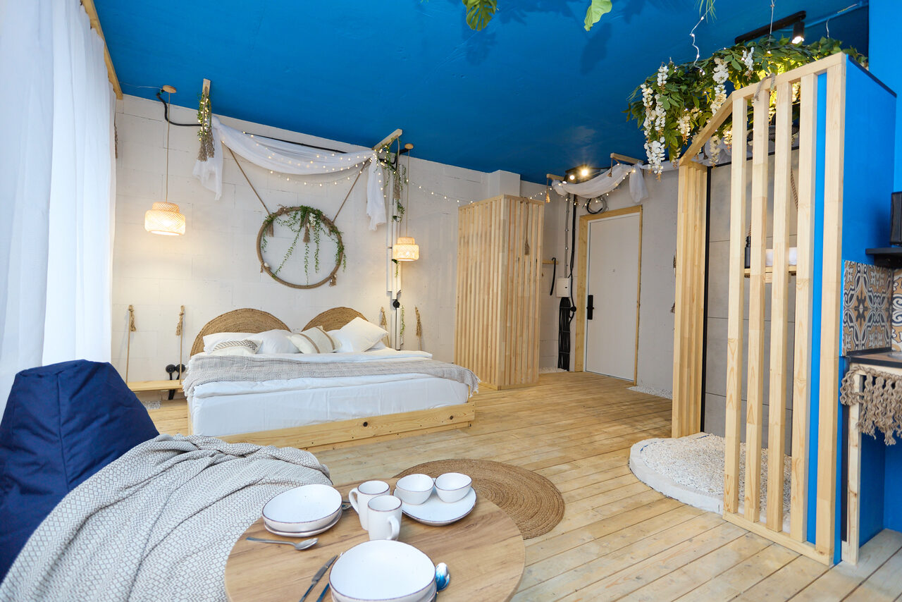 Арендовать Лофт Бали с фото зоной на 23 этаже с видом на Москва-реку. Просторная комната с синим потолком, деревянным полом и светлыми стенами. Двуспальная кровать с плетеными изголовьями. Деревянный стол с посудой, подвесные светильники, зеленые растения, диван, белый текстиль, деревянные перегородки, декоративные элементы на стенах.