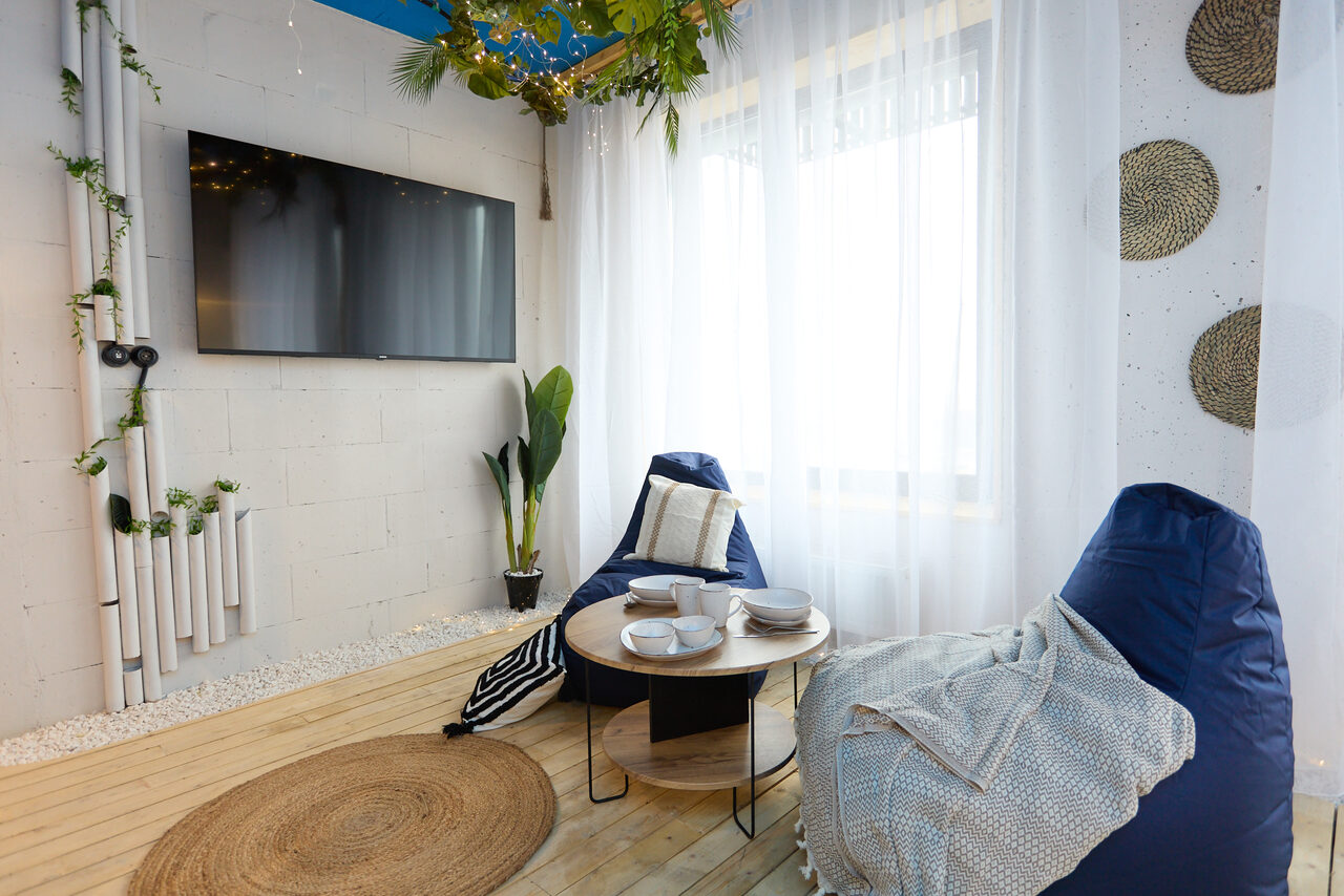 Арендовать Лофт Бали с фото зоной на 23 этаже с видом на Москва-реку. Комната с деревянным полом и белыми кирпичными стенами, телевизор на стене, два мягких кресла-мешка, круглые столики с посудой, большое окно с прозрачными шторами, плетёные декоративные элементы на стене, зеленые растения, светлое и уютное освещение.