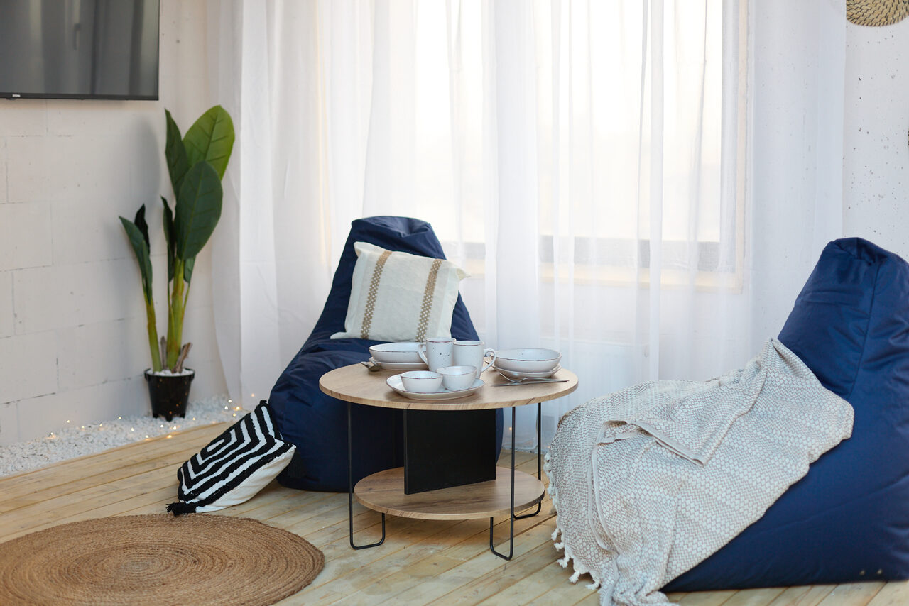 Арендовать Лофт Бали с фото зоной на 23 этаже с видом на Москва-реку. Комната с деревянным полом, двумя синими креслами-мешками, круглым деревянным столиком, несколькими чашками, пледами и подушками. Белые прозрачные занавески, комнатное растение в углу, настенное освещение.