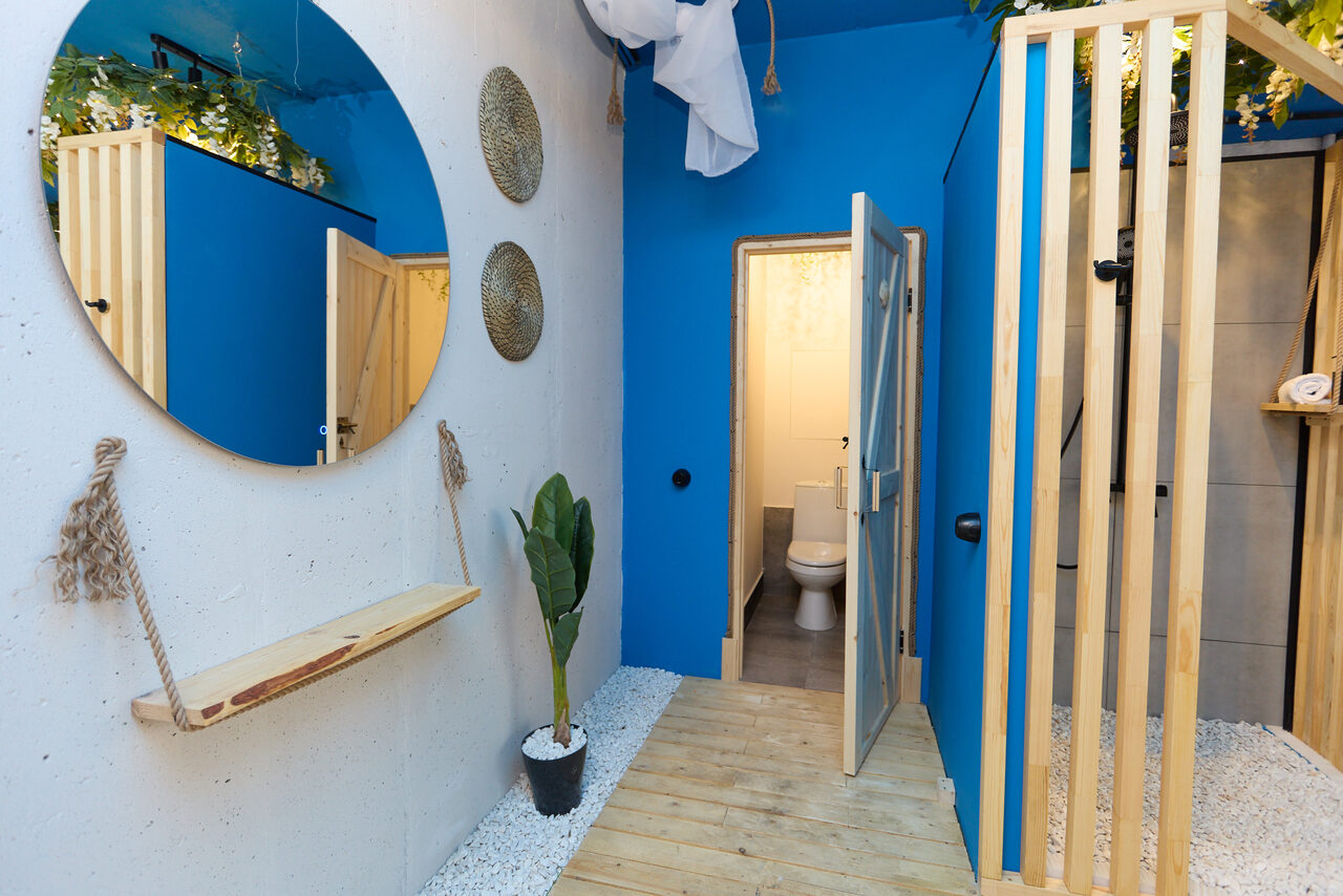 Арендовать Лофт Бали с фото зоной на 23 этаже с видом на Москва-реку. Круглое зеркало на белой стене, подвесная полка, декоративные корзины, синий и белый цвета, деревянный пол, открытая дверь в туалетную комнату, стены с элементами деревообработки, растение в горшке, белый гравий.