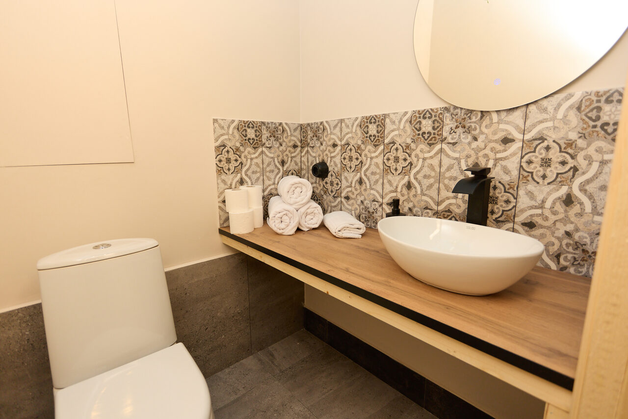 Арендовать Лофт Бали с фото зоной на 23 этаже с видом на Москва-реку. Туалет с унитазом, настенным зеркалом и раковиной на деревянной столешнице. Узоры на плитке на стене, небольшое настенное зеркало круглой формы, мраморные полы. Бумажные полотенца и туалетная бумага на столешнице. Светлые стены и стены из керамогранита.