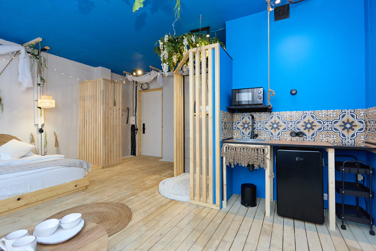 Арендовать Лофт Бали с фото зоной на 23 этаже с видом на Москва-реку. Студия с деревянным полом и синим потолком. Кровать слева, кухонный уголок справа с синим акцентом и орнаментальной плиткой, микроволновкой, маленьким холодильником. Деревянные декоративные элементы, светлая мебель, искусственные растения, мягкое освещение.