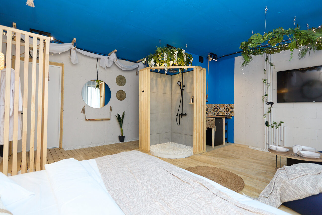 Арендовать Лофт Бали с фото зоной на 23 этаже с видом на Москва-реку. Спальня-студия с деревянным полом, кроватью и зоной душа. Голубые и белые стены, потолок ярко-синего цвета. Зеркало круглой формы на стене, несколько круглых декоративных элементов, зелёные растения, телевизор. Светильники и декор на потолке.