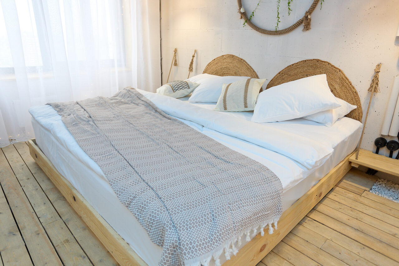 Арендовать Лофт Бали с фото зоной на 23 этаже с видом на Москва-реку. Спальня с двуспальной кроватью на деревянном каркасе. Светлое покрывало, подушки. Декор на стене с круглыми элементами. Деревянный пол, светлые шторы на окне. Белая стена с легким фактурным рисунком. Нейтральная цветовая гамма.