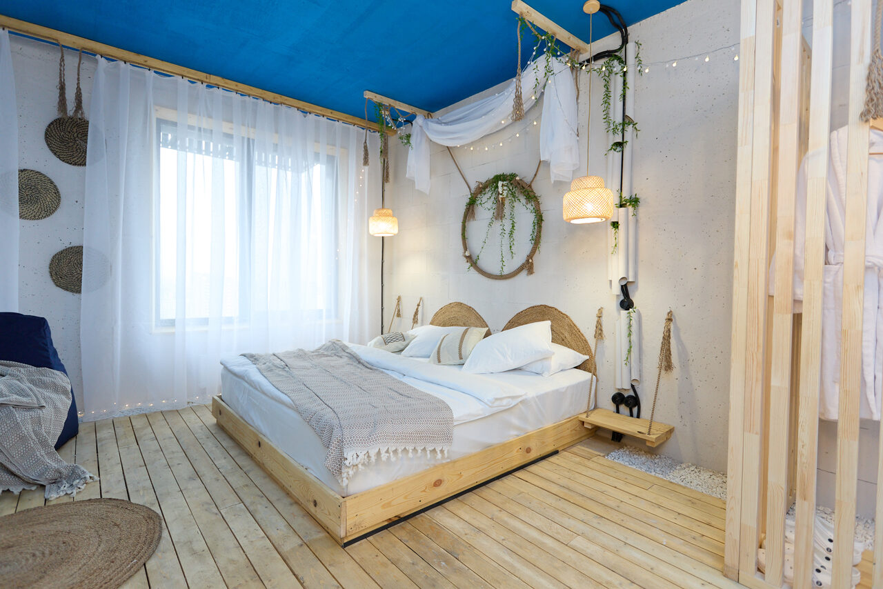 Арендовать Лофт Бали с фото зоной на 23 этаже с видом на Москва-реку. Спальня с деревянным настилом и большой кроватью. Светлые стены, голубой потолок, белые шторы на окне. Подвесные светильники, украшения из лозы и зелени. Подушки и плед на кровати. Натуральные материалы, скандинавский стиль.