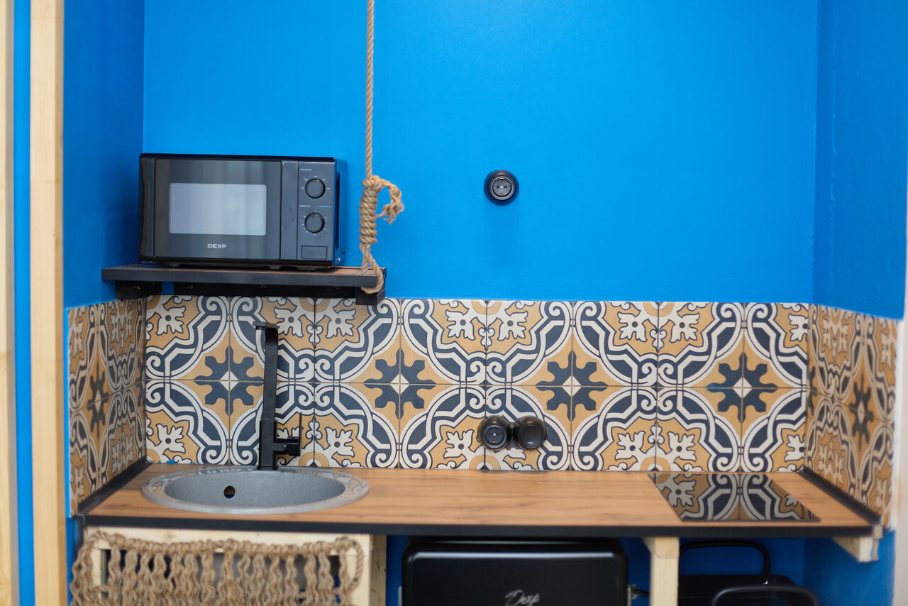 Арендовать Лофт Бали с фото зоной на 23 этаже с видом на Москва-реку. Кухонная зона с синими стенами и узорчатым плиточным фартуком. Имеется деревянная столешница с круглой раковиной, подвесная плита и черная микроволновая печь на полке. Рядом плетеная корзина и розетка.