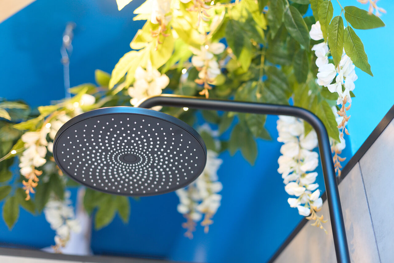 Арендовать Лофт Бали с фото зоной на 23 этаже с видом на Москва-реку. Душевая зона с круглой черной лейкой на изогнутом держателе, декоративные зелёные листья и белые цветы сверху, стены голубого цвета.