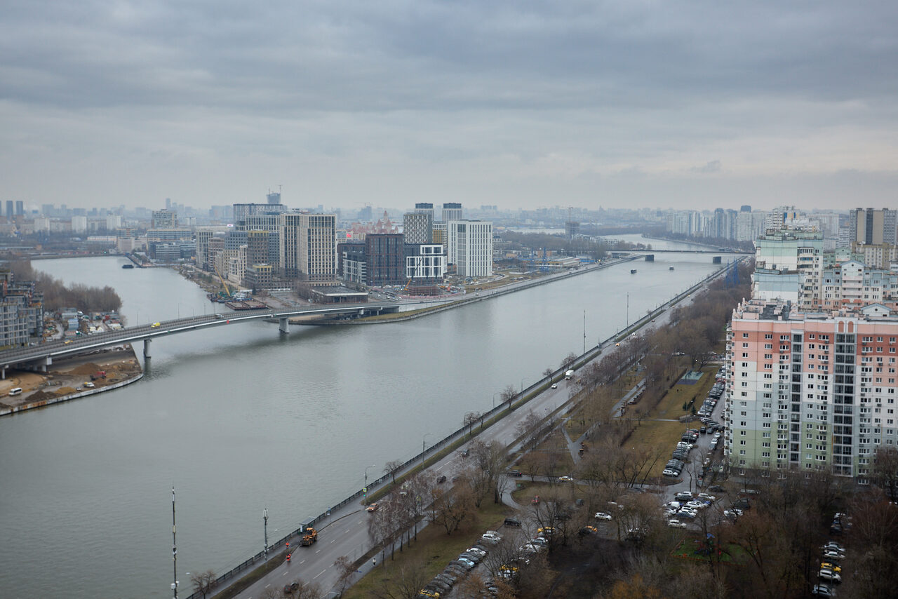 Арендовать Лофт Бали с фото зоной на 23 этаже с видом на Москва-реку. Вид на город с высоты, река проходит между зданиями, моста соединяет берега. Высотные жилые и коммерческие здания, серый облачный день, дорога вдоль набережной, автомобили припаркованы вдоль улицы.