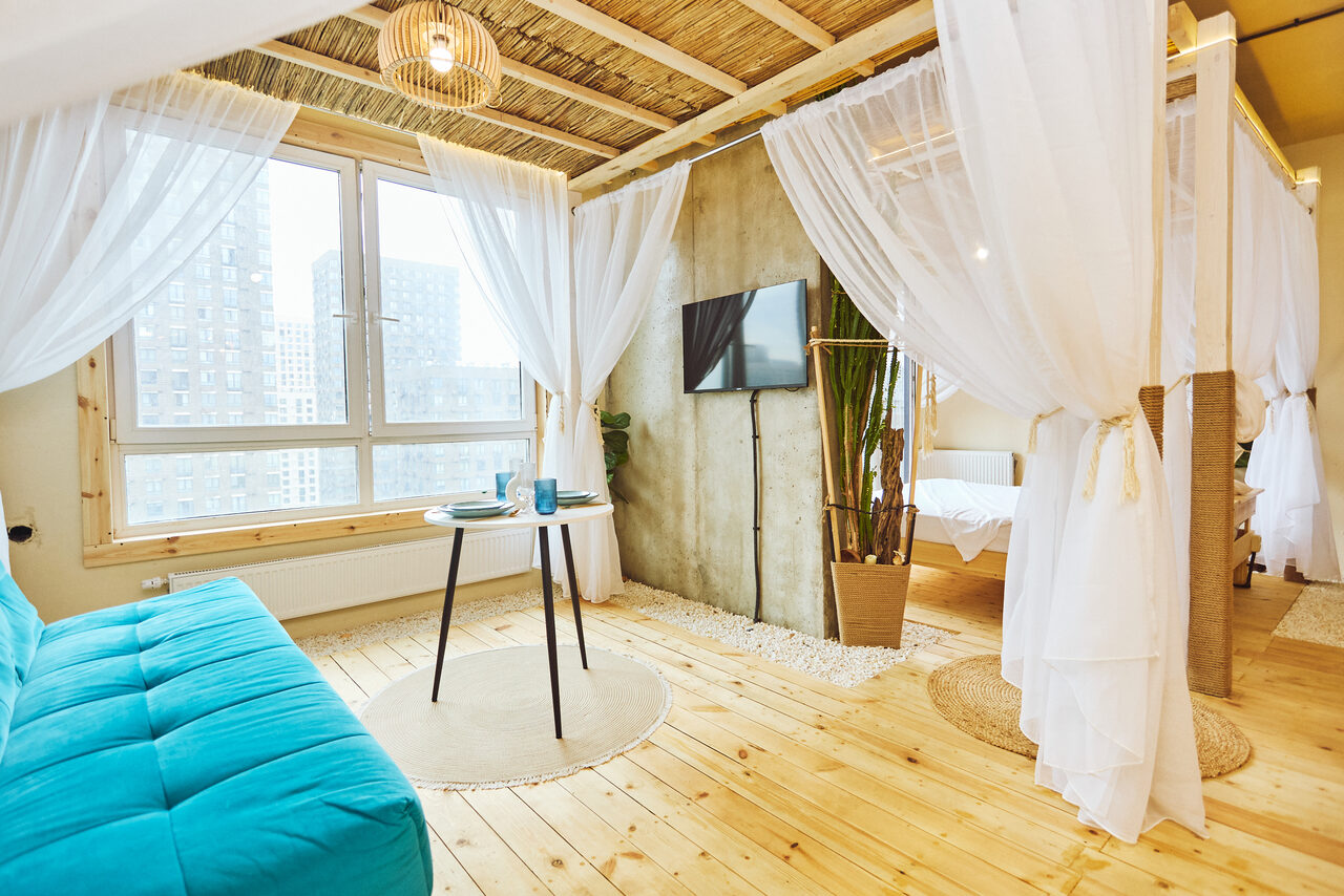 Арендовать Тропический лофт с бунгало, баром и французским балконом. Просторное светлое помещение с деревянным полом и потолком. Большое окно, разделённое занавесками на зоны. Бирюзовый диван, круглый стол с двумя стаканами, телевизор на стене, крупное растение в горшке. Светлая ткань занавесок, светильник на потолке.