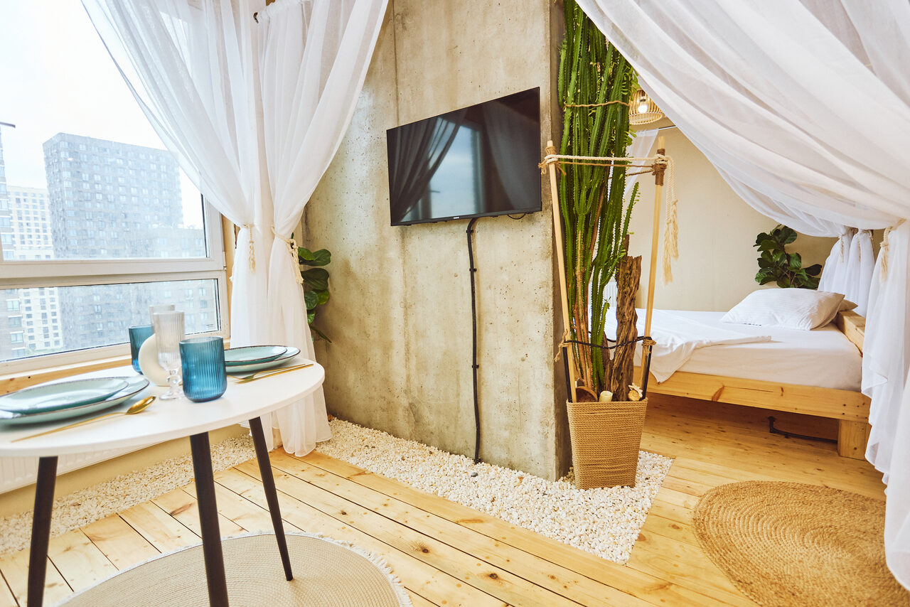 Арендовать Тропический лофт с бунгало, баром и французским балконом. Просторная комната с деревянным полом и бетонной стеной. Круглый белый стол с двумя приборами, телевизор на стене, кровать с белым покрывалом. Растения в горшке, белые занавески, мягкое дневное освещение, большое окно, коврик из натурального материала.