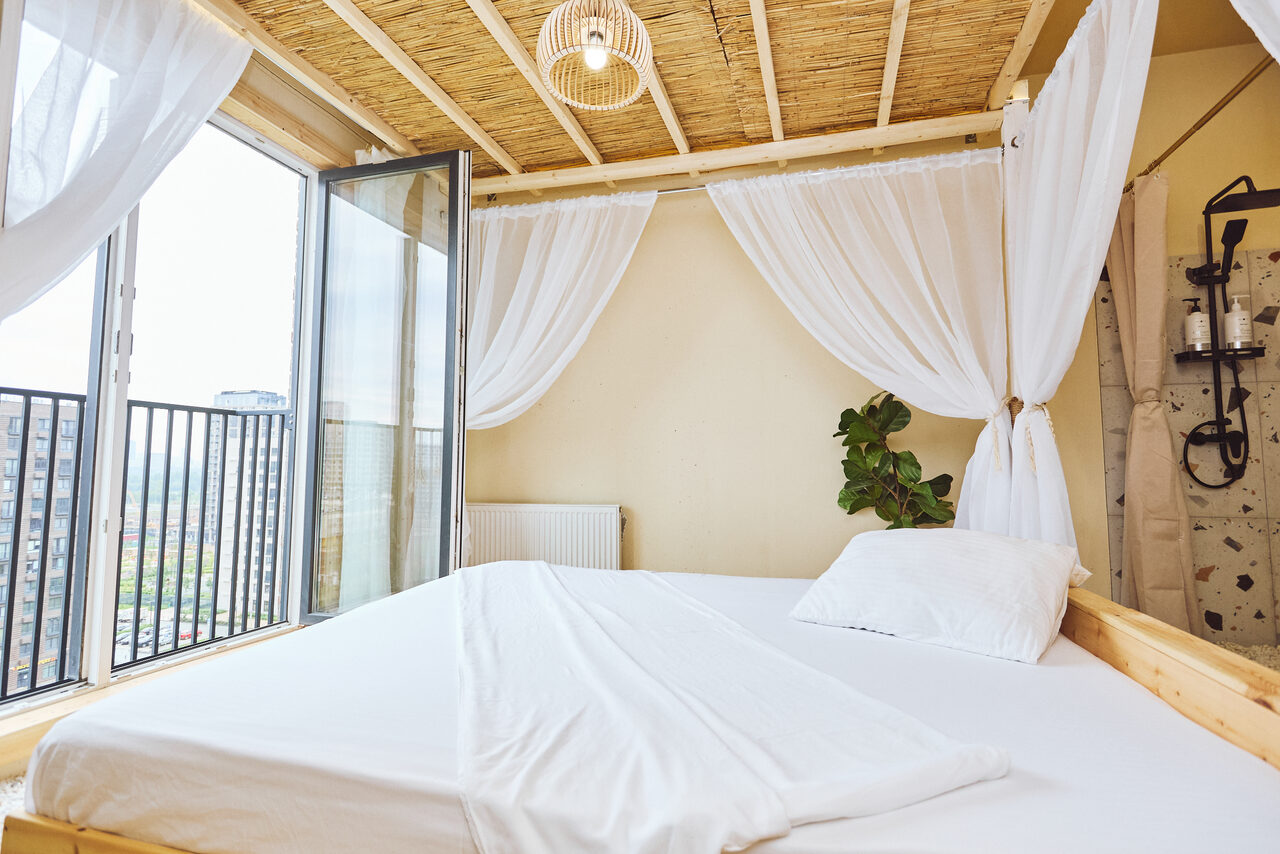 Арендовать Тропический лофт с бунгало, баром и французским балконом. Спальня с двуспальной кроватью, белыми простынями и подушками. Над кроватью белые полупрозрачные шторы. Открытые двери на балкон с черными металлическими перилами. Пол светлый, стены бежевые. Потолок из дерева с декоративным светильником.