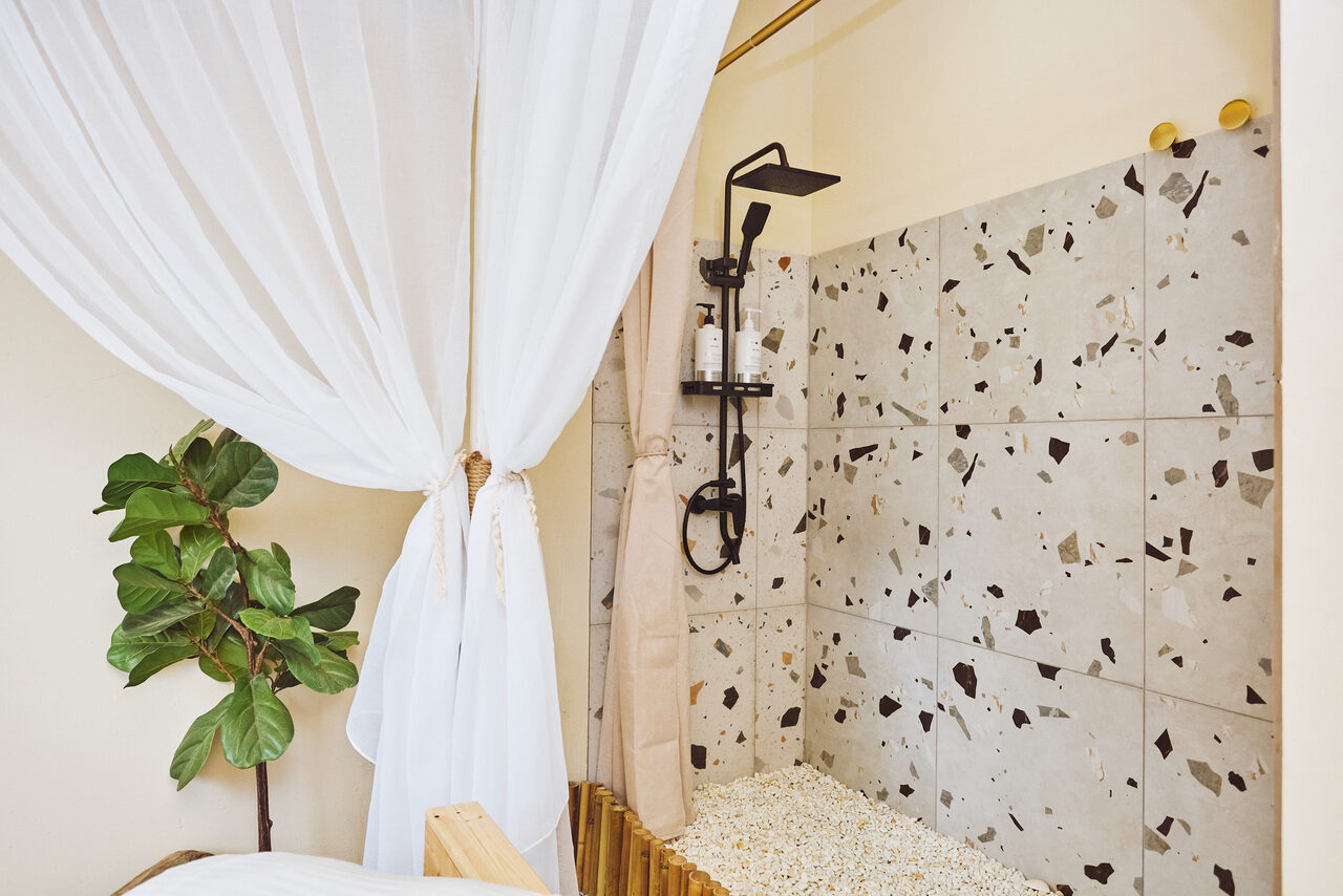 Арендовать Тропический лофт с бунгало, баром и французским балконом. Душевая зона с терраццо плиткой, черный душевой комплект, прозрачные диспенсеры для шампуня. Светлый текстильный занавес, пастельные стены. Зеленое растение в углу. Пастельные и нейтральные тона.