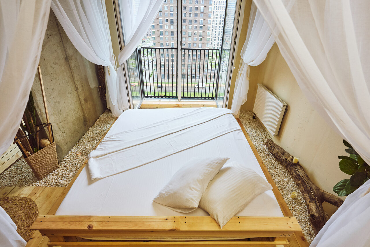 Арендовать Тропический лофт с бунгало, баром и французским балконом. Спальня с большой кроватью на деревянном каркасе, украшенная белыми занавесками. Пол из светлого дерева с ковриком. Окно с балконом, вид на город. Стена из бетона с растениями и декором из дерева. Радиатор на стене.