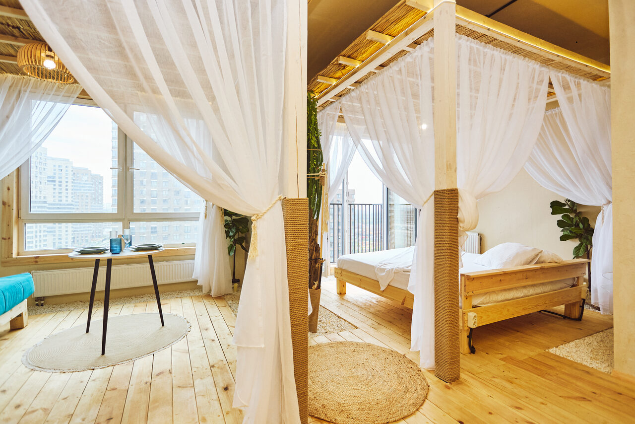 Арендовать Тропический лофт с бунгало, баром и французским балконом. Спальня-студия с деревянным полом, двумя кроватями с балдахинами, прозрачными шторами. Круглый стол с книгой и кружкой на коврике. Окно с видом на небоскрёбы, мягкое дневное освещение. Светлые натуральные тона, зелёные растения.