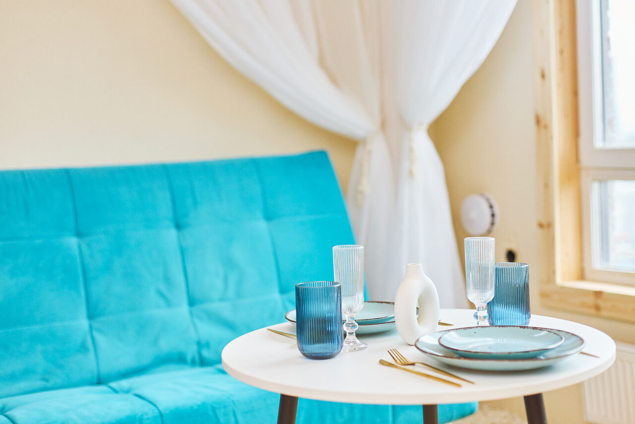 Арендовать Тропический лофт с бунгало, баром и французским балконом. Гостиная, диван с бирюзовой обивкой, круглый стол с синей и белой посудой, золотистые столовые приборы. Белые прозрачные шторы, деревянные элементы интерьера, светлые стены, окно с двойной рамой.