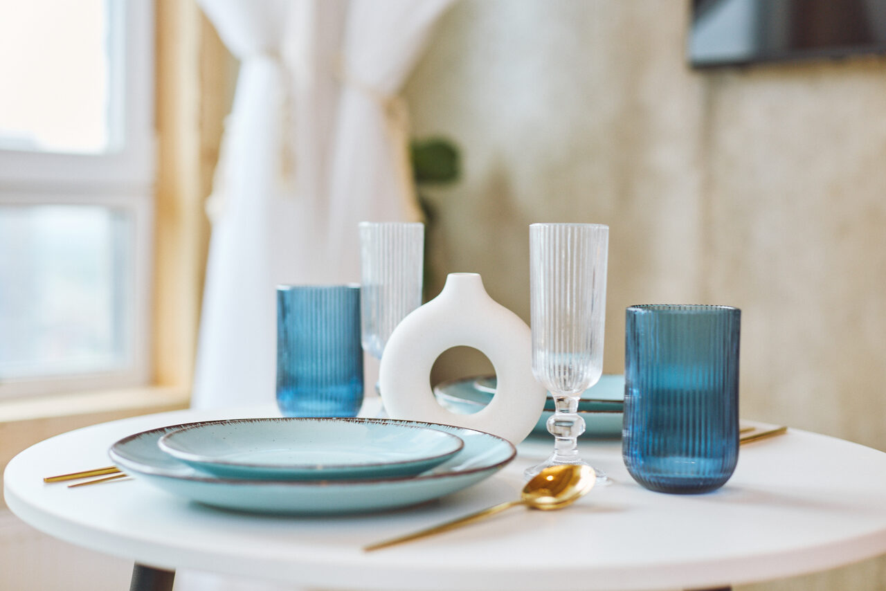 Арендовать Тропический лофт с бунгало, баром и французским балконом. Белый круглый стол с сервировкой: тарелки голубого цвета, золотые столовые приборы, стеклянные бокалы и синяя посуда, белая ваза с отверстием. Светло-бежевые стены, окно с белой шторой на заднем плане. Освещение естественное.