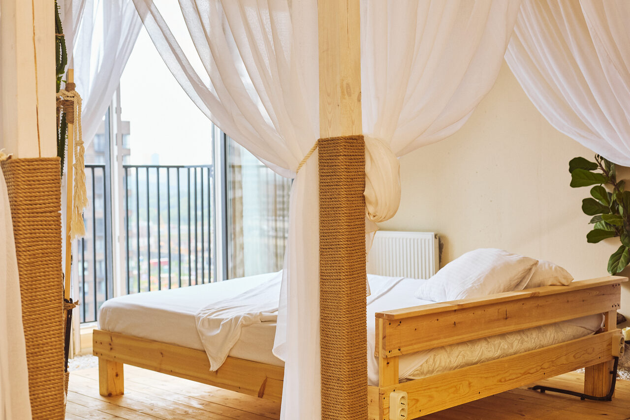 Арендовать Тропический лофт с бунгало, баром и французским балконом. Кровать с балдахином в светлой комнате, деревянный пол, белые прозрачные занавески, выход на балкон с металлическими перилами, растительный декор, светлые стены, подушки и одеяло белого цвета, радиатор отопления у стены.