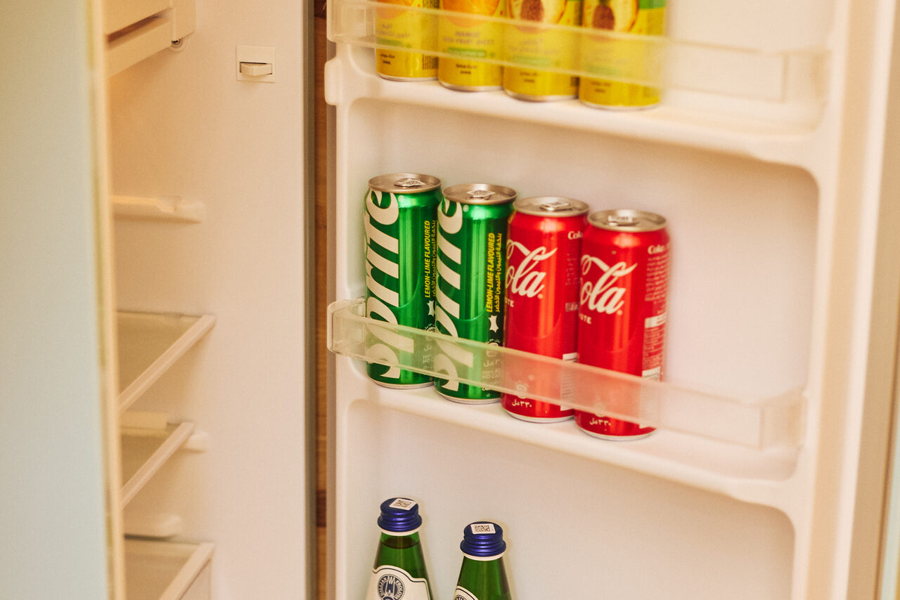 Арендовать Тропический лофт с бунгало, баром и французским балконом. Открытый холодильник с напитками. На дверце расположены банки с газировкой, в основном зелёного и красного цветов. На полках видны стеклянные бутылки. Холодильник белого цвета, с пустыми основными полками. Освещение тёплое.