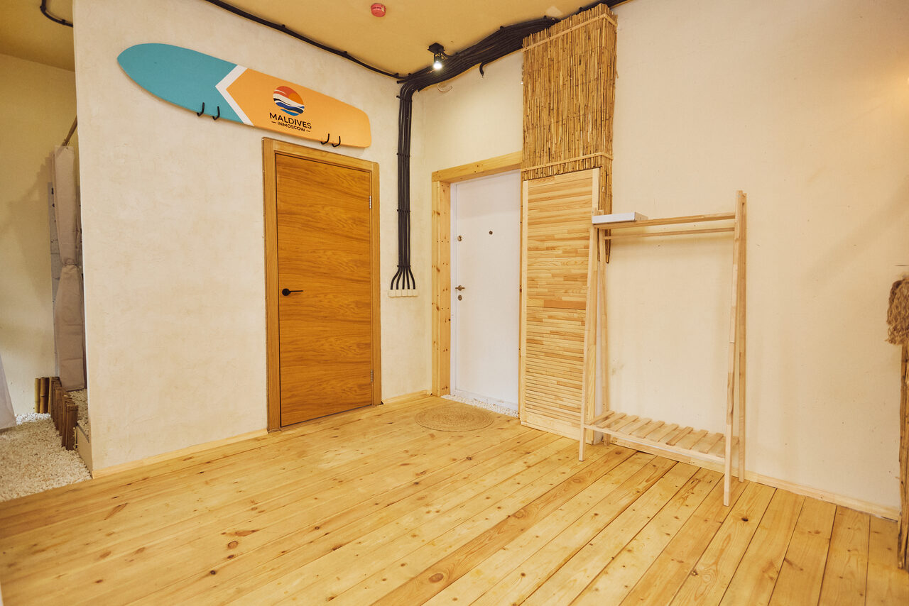 Арендовать Тропический лофт с бунгало, баром и французским балконом. Комната с деревянным полом, светлыми стенами и несколькими дверьми. На стене висит декор в виде доски для серфинга, рядом деревянная вешалка без одежды. Потолок бежевый с черными трубами. Отделка из бамбуковых панелей.