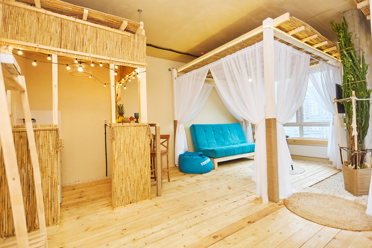 Арендовать Тропический лофт с бунгало, баром и французским балконом. Пространство с деревянным полом, перегородками из бамбука, зоной отдыха с бирюзовым диваном и креслом-мешком, белыми занавесками. Барная стойка с высокими стульями, гирлянда с лампами, текстильные ковры, большие растения, окно с белой рамой.