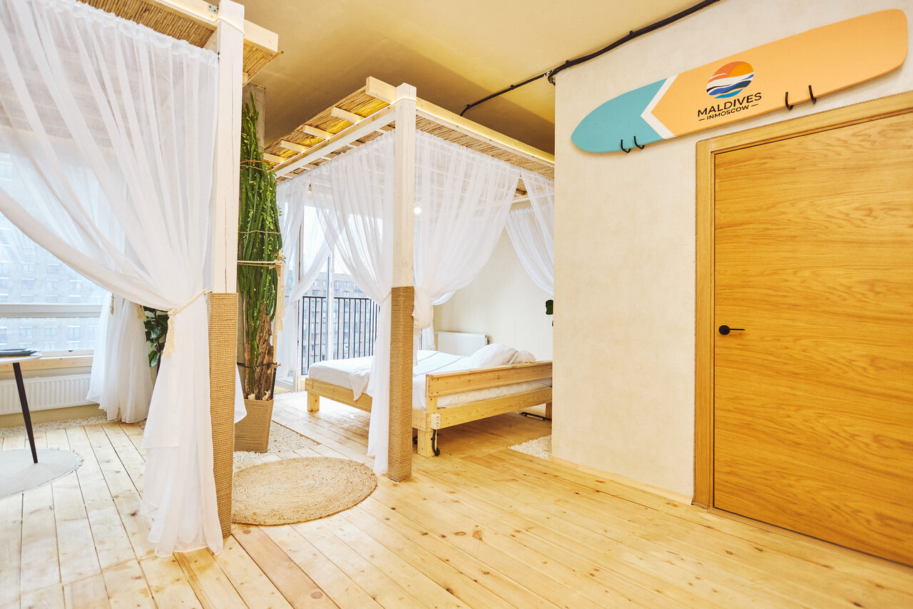 Арендовать Тропический лофт с бунгало, баром и французским балконом. Спальня с балдахином, деревянная кровать, белые прозрачные шторы, деревянный пол, светлая отделка стен. На стене декор в виде стилизованной доски для серфинга, круглый ковер, крупное растение, стеклянный оконный проем.