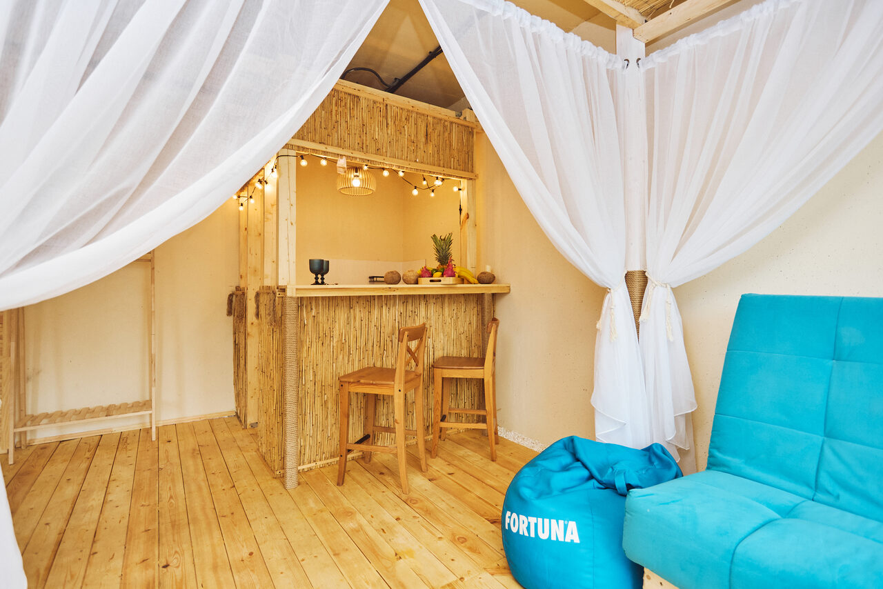Арендовать Тропический лофт с бунгало, баром и французским балконом. Пространство с деревянным полом и барной стойкой из бамбука. Две деревянные табуретки, белые полупрозрачные шторы, голубое кресло и мешок-кресло с надписью "Fortuna". Подвесное освещение с лампочками и фруктами на стойке.