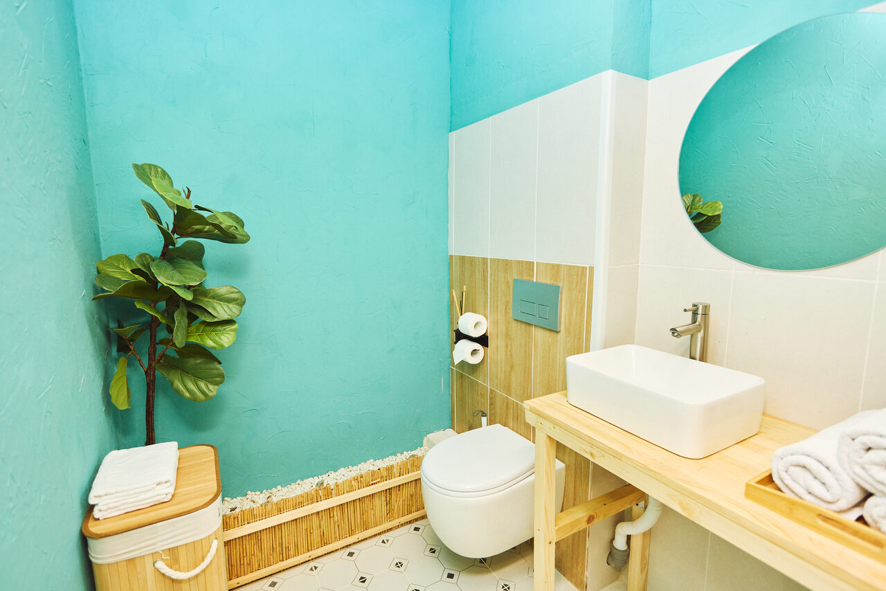 Арендовать Тропический лофт с бунгало, баром и французским балконом. Ванная комната с бирюзовыми стенами и белой плиткой, круглое зеркало, деревянная раковина с белой мойкой, полотенца, унитаз, рулоны туалетной бумаги, декоративное растение, корзина.