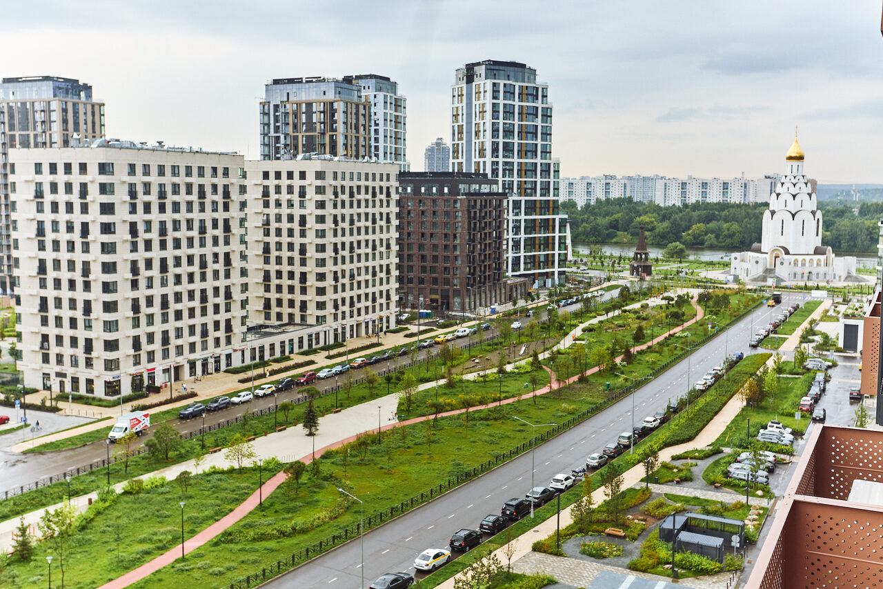 Арендовать Тропический лофт с бунгало, баром и французским балконом. Современные многоэтажные жилые здания с разнообразным дизайном фасадов вдоль аллеи с зелёными насаждениями, машин на парковке, церковь с золотым куполом, пешеходная зона и газоны между дорогами, частично облачное небо.