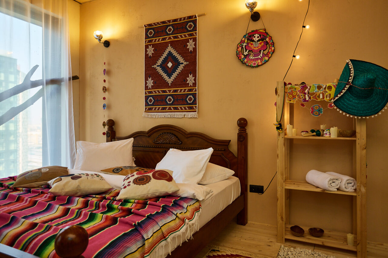 Арендовать ⁠Лофт в Мексиканском стиле для костюмированных вечеринок. Спальня с деревянной кроватью, покрытой разноцветным пледом и подушками. Стена украшена текстильным панно и декоративными тарелками. Рядом деревянный стеллаж с полотенцами. Стены светло-коричневые, на полу светлая древесина. Естественное и точечное освещение.