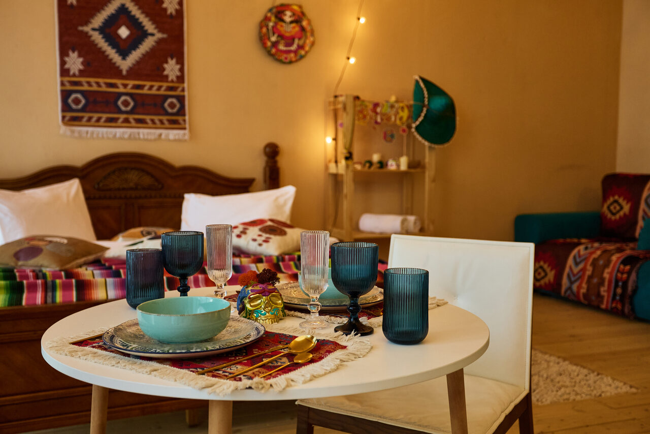 Арендовать ⁠Лофт в Мексиканском стиле для костюмированных вечеринок. Круглый белый стол с разноцветной посудой, накрытый тканью с этническим узором, рядом белый стул. На фоне кровать с подушками и покрывалом, декорированные стены, светящаяся гирлянда, полка, рядом синие и красные элементы декора, ковёр на полу.