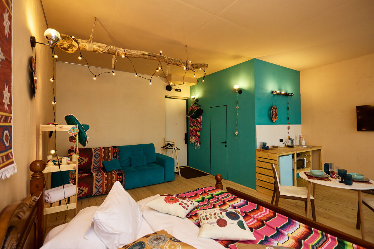 Арендовать ⁠Лофт в Мексиканском стиле для костюмированных вечеринок. Комната с кроватью, диваном и обеденным столом. Яркие текстильные элементы. Голубая акцентная стена с дверью. Настенная полка и стойка с посудой. Деревянный пол и кровать. Лампочки на проводе, подсветка над столиком. Светлая отделка, декоративные элементы.