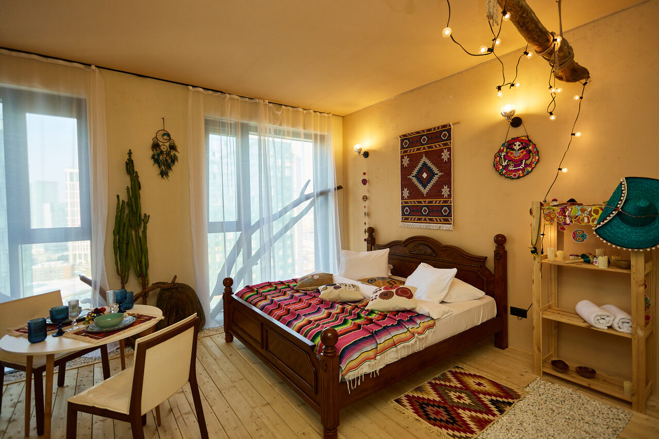 Арендовать ⁠Лофт в Мексиканском стиле для костюмированных вечеринок. Спальня с деревянным полом, двуспальная кровать с ярким покрывалом, настенные декорации, стол с двумя стульями, открытая деревянная полка с предметами, большие окна с полупрозрачными шторами, растения, подвесное освещение на ветке.