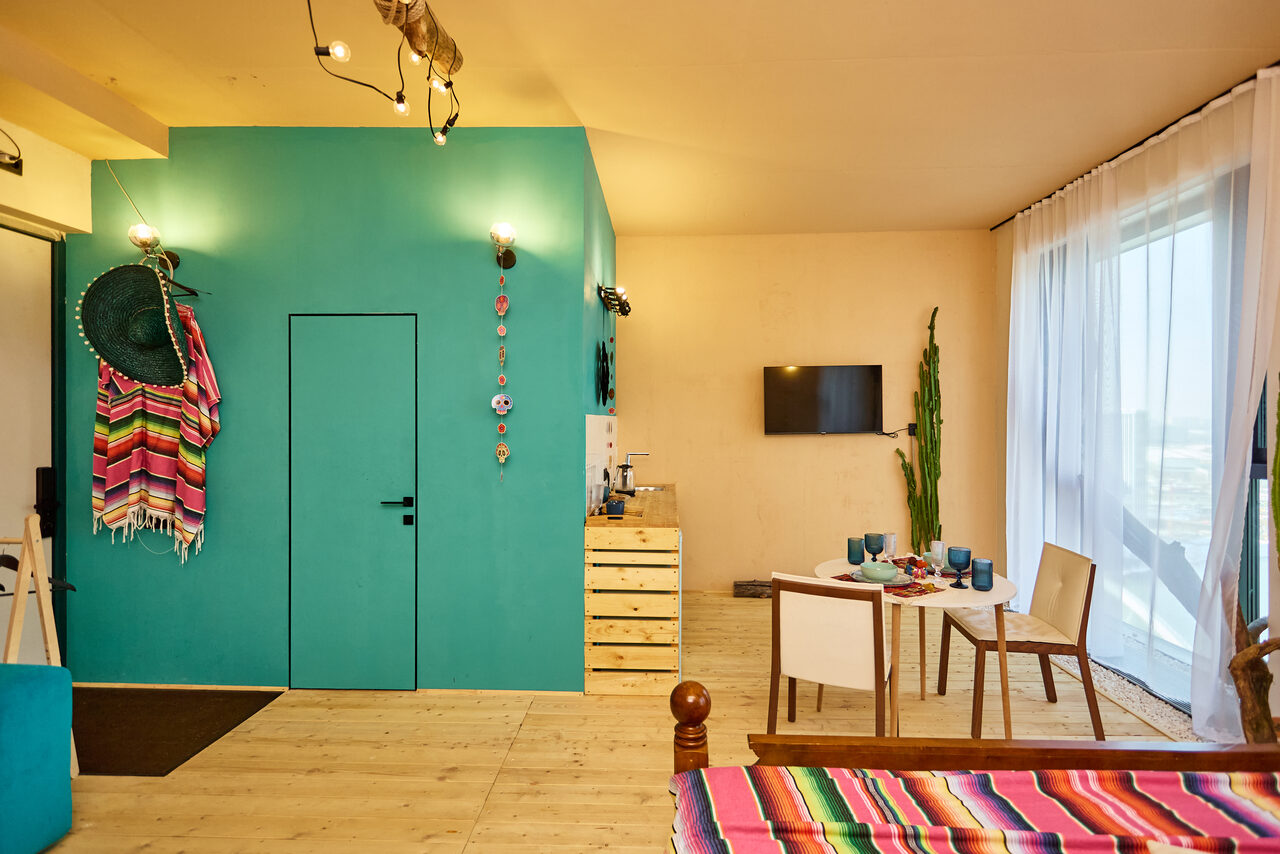 Арендовать ⁠Лофт в Мексиканском стиле для костюмированных вечеринок. Гостиная с ярко-голубыми и бежевыми стенами, деревянный пол, стол с посудой и двумя стульями, ТВ на стене, декоративные элементы, кактус, цветные покрывала и шляпы, мягкое освещение, большие окна с белыми шторами.