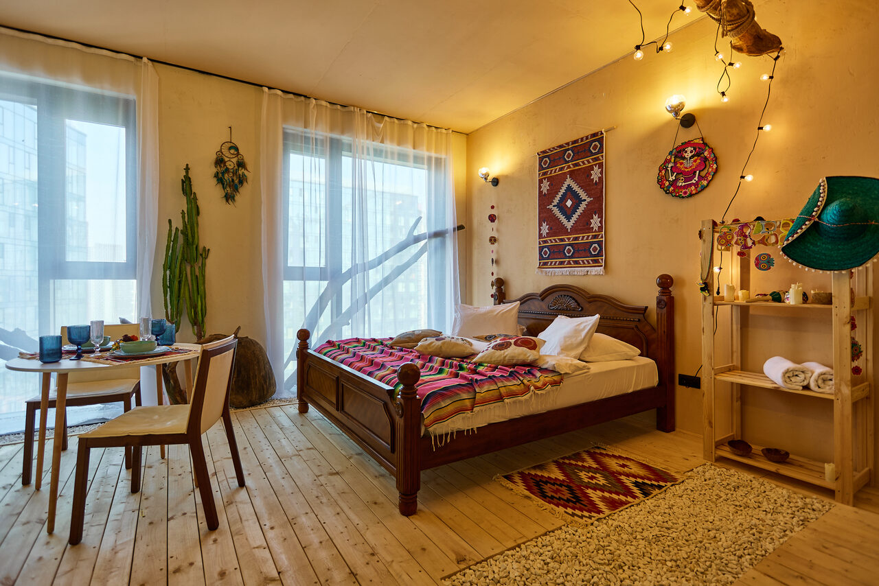 Арендовать ⁠Лофт в Мексиканском стиле для костюмированных вечеринок. Спальня с деревянным полом, большая кровать с ярким покрывалом, рядом круглый обеденный стол с двумя стульями, окно с прозрачными шторами, деревянный стеллаж с декоративными элементами, стены украшены текстильными панно, мягкое рассеянное освещение.