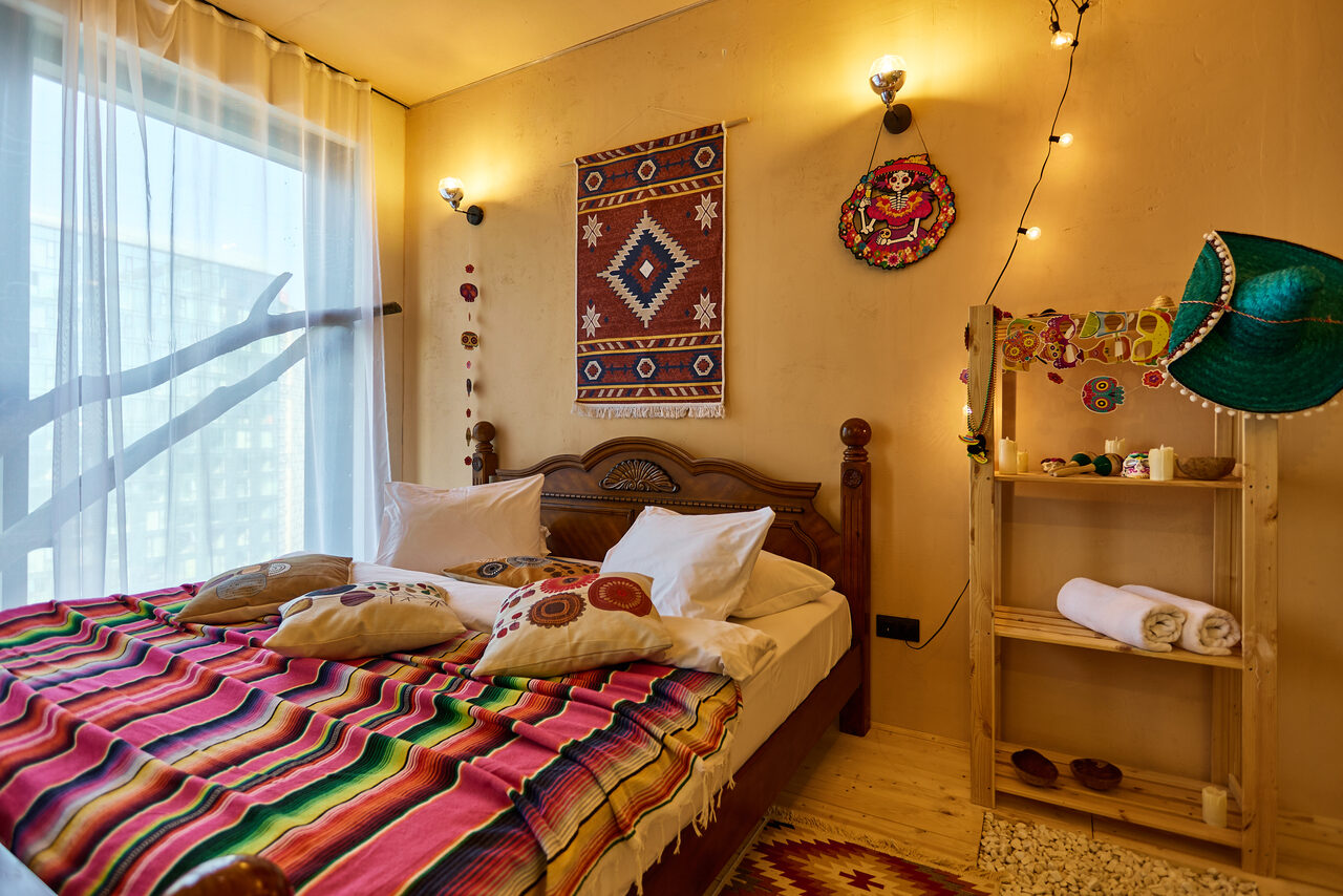 Арендовать ⁠Лофт в Мексиканском стиле для костюмированных вечеринок. Спальня с деревянной кроватью, цветастым покрывалом и подушками. Стена украшена ковриком и декором. Полка с полотенцами и украшениями. Большое окно с прозрачной шторой. Теплое освещение от настенных светильников. Светлые стены и пол.