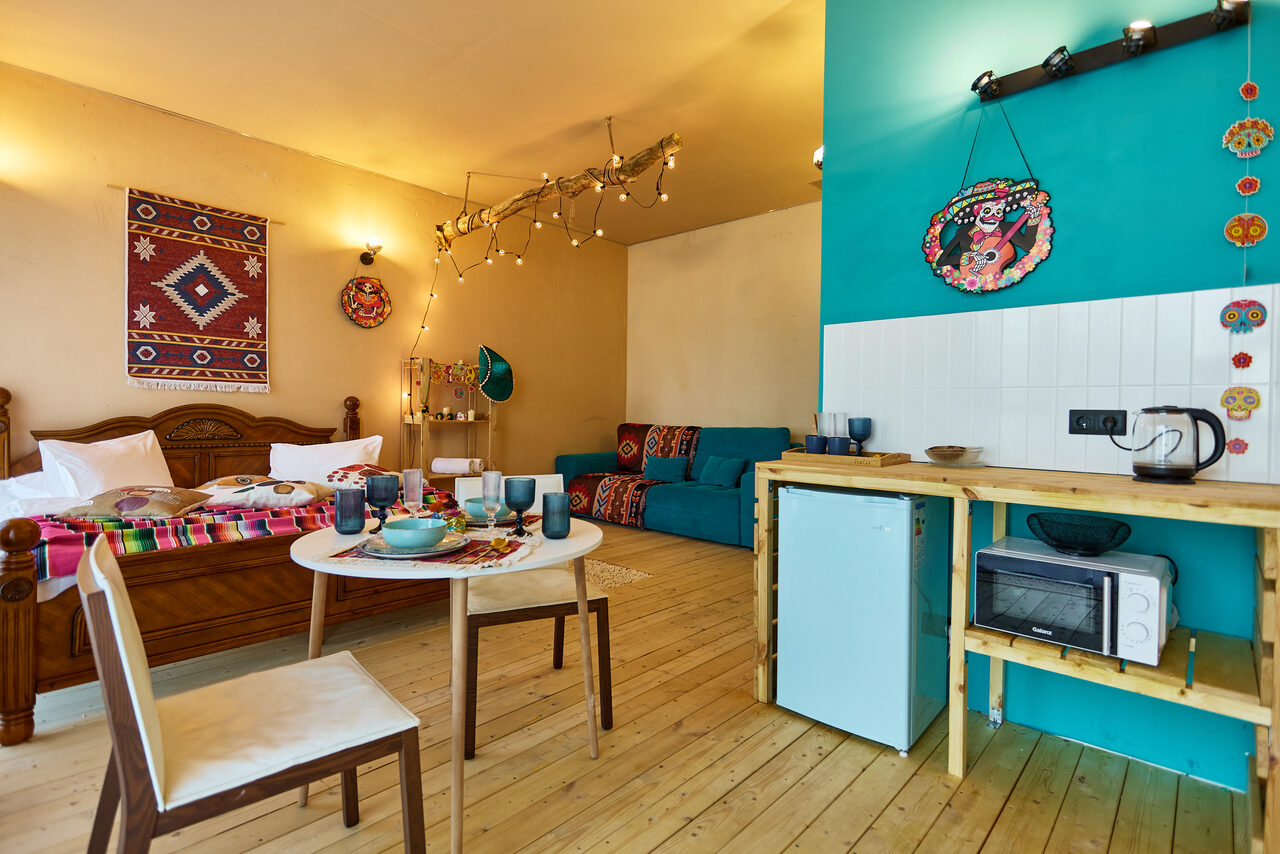 Арендовать ⁠Лофт в Мексиканском стиле для костюмированных вечеринок. Спальня-гостиная с деревянным полом, двуспальной кроватью и диваном. Кухонный уголок с микроволновкой, плитой и мини-холодильником. Стены светлые и бирюзовые. Декор на стенах, гирлянда и настенные светильники. деревянный стол с двумя стульями.