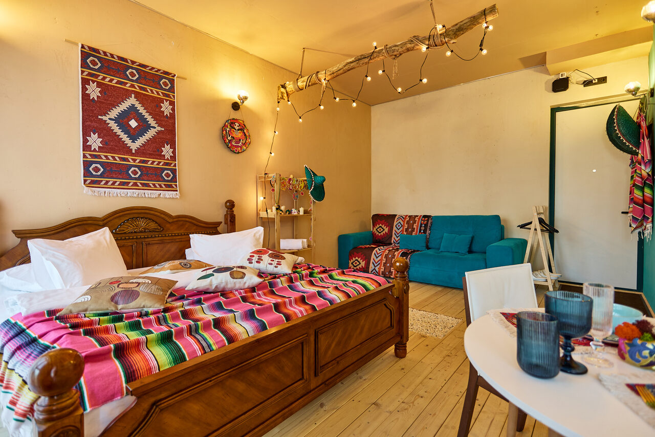 Арендовать ⁠Лофт в Мексиканском стиле для костюмированных вечеринок. Спальня с деревянной кроватью, покрытой ярким полосатым покрывалом. Синий диван, деревянный пол, белый круглый стол с посудой. Стена украшена ковром с этническим узором. Подвесные лампочки на ветке. Светлые стены.