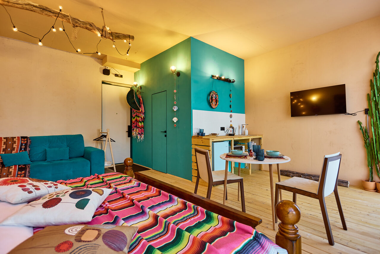 Арендовать ⁠Лофт в Мексиканском стиле для костюмированных вечеринок. Гостевая комната с яркой кроватью, деревянным полом, голубым диваном и круглым столом с двумя стульями. Оборудована настенным телевизором и декоративными элементами. Комната освещена светодиодными лампами, стены окрашены в бежевый и бирюзовый.