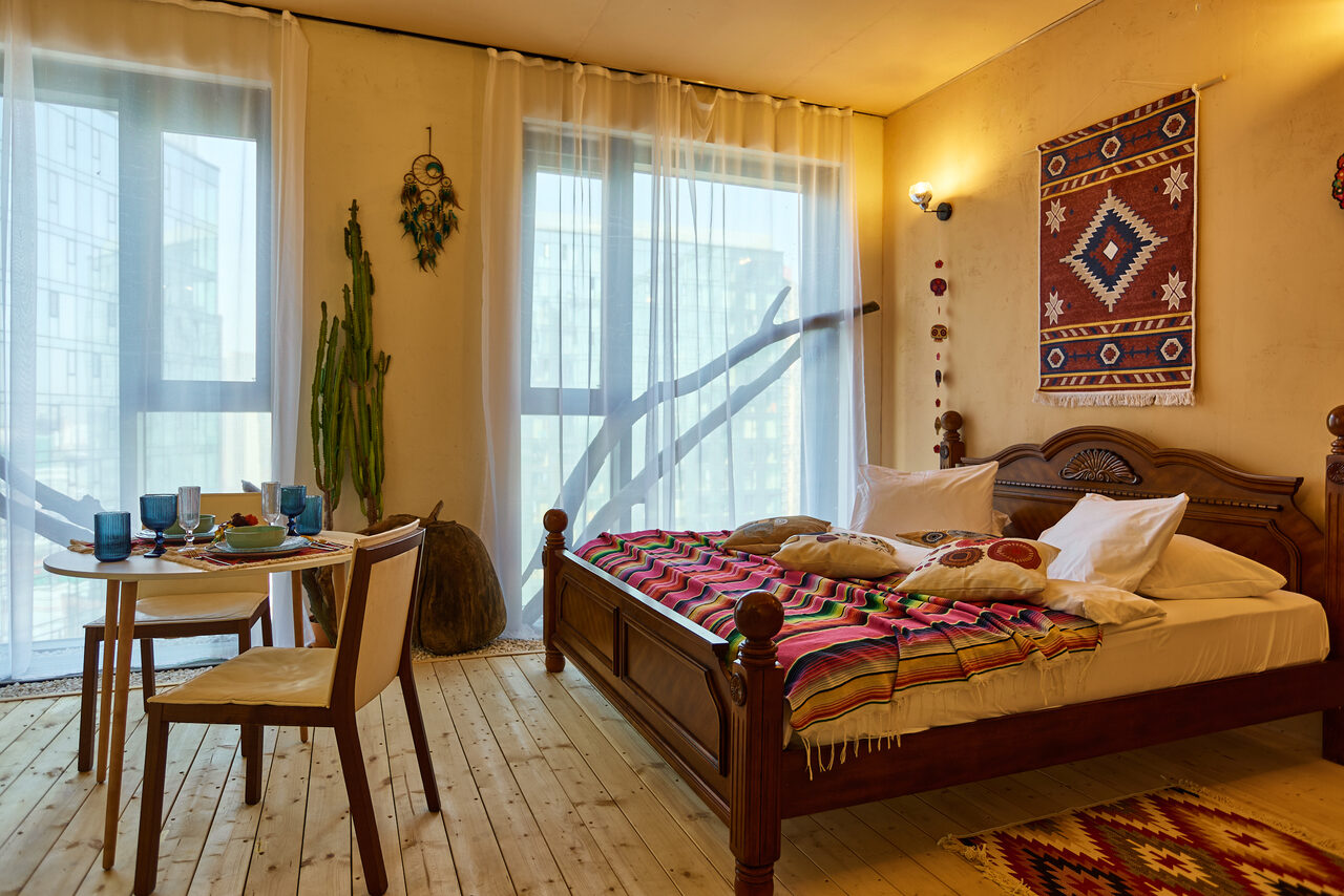 Арендовать ⁠Лофт в Мексиканском стиле для костюмированных вечеринок. Спальня с деревянным полом, двуспальной кроватью, декорированной цветными текстилями и подушками. Стол с посудой и двумя стульями. Светлые шторы на окнах, настенное украшение и кактус. Светлая стена с ковром и бра.