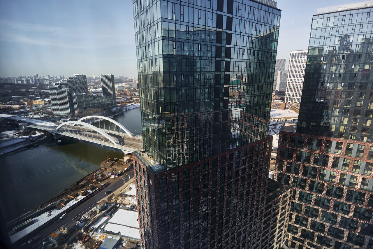 Арендовать ⁠Лофт в Мексиканском стиле для костюмированных вечеринок. Вид из окна на городскую панораму с двумя высокими стеклянными зданиями, рекой и белым арочным мостом. На заднем плане виднеются другие многоэтажные здания. Цвета: синий, белый, коричневый.