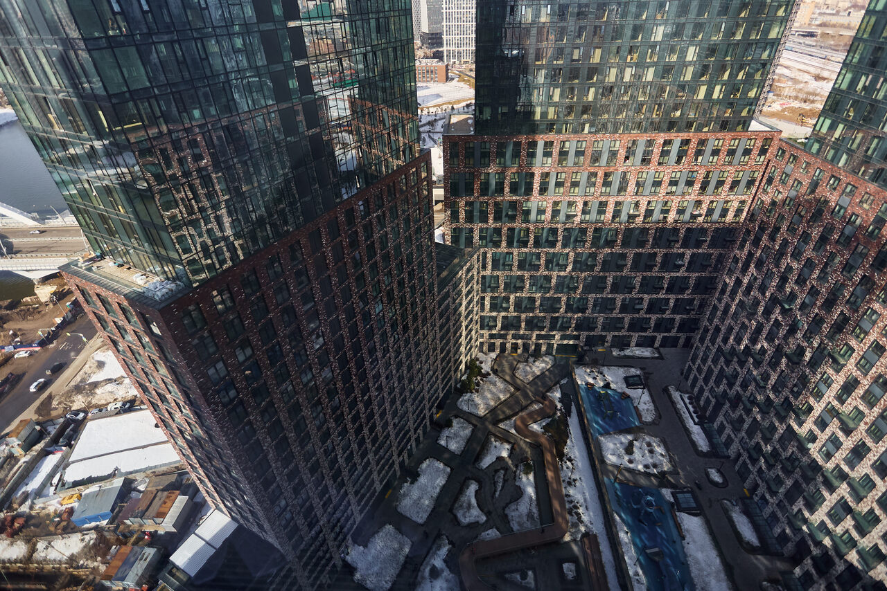 Арендовать ⁠Лофт в Мексиканском стиле для костюмированных вечеринок. Высотные здания с многоэтажным фасадом из стекла и кирпича, образуют внутренний двор. На уровне земли видны тротуары с остатками снега. Вдалеке виден небольшой участок реки и окружающая городская застройка. Освещение естественное, солнечное.