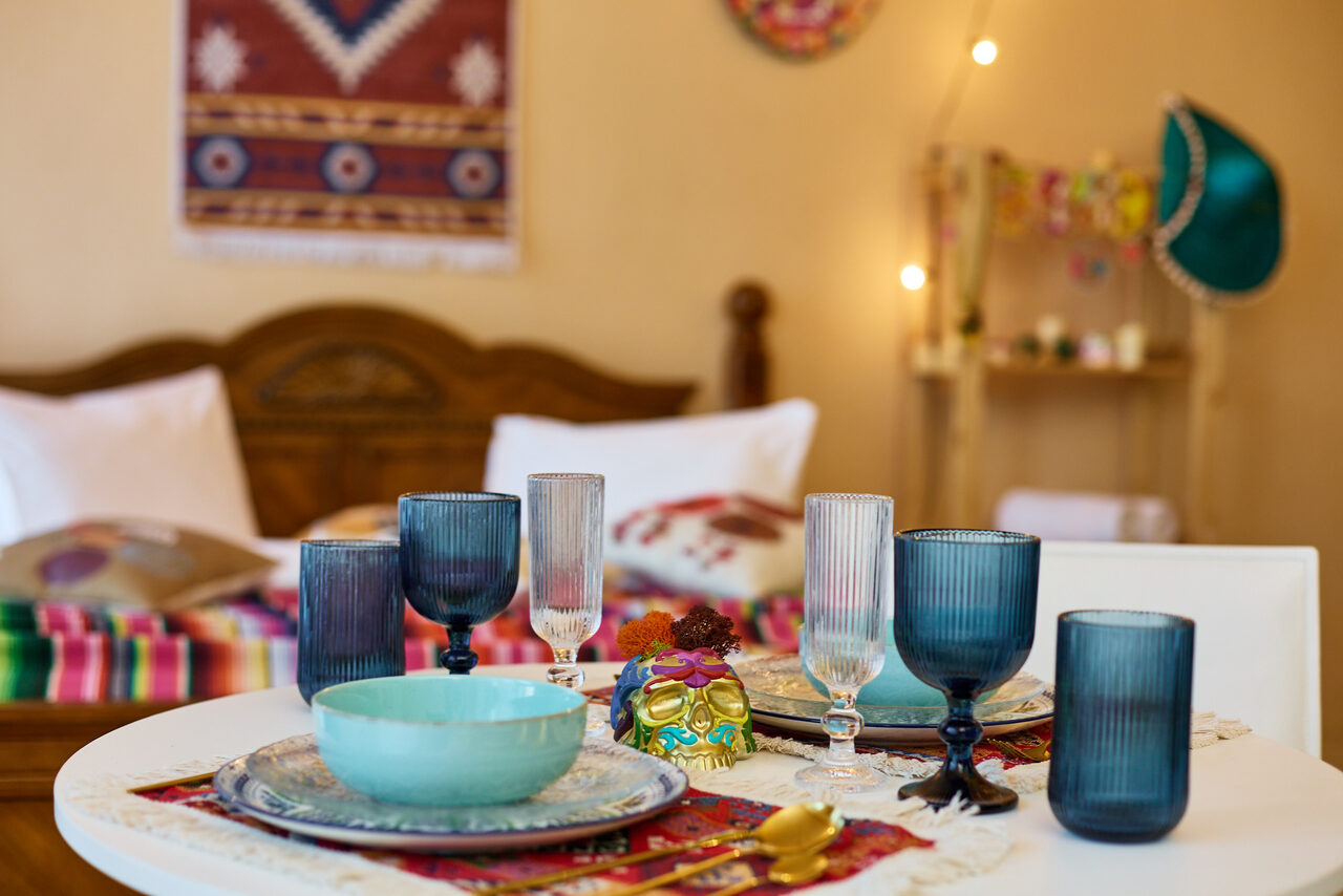 Арендовать ⁠Лофт в Мексиканском стиле для костюмированных вечеринок. Круглый стол с сервировкой посуды и бокалов в разных цветах, декорированный скатертью с узорами. На заднем плане кровать с подушками, покрытая цветным пледом. На стене гобелен, мягкое освещение от фонариков. Цвета теплые и яркие.