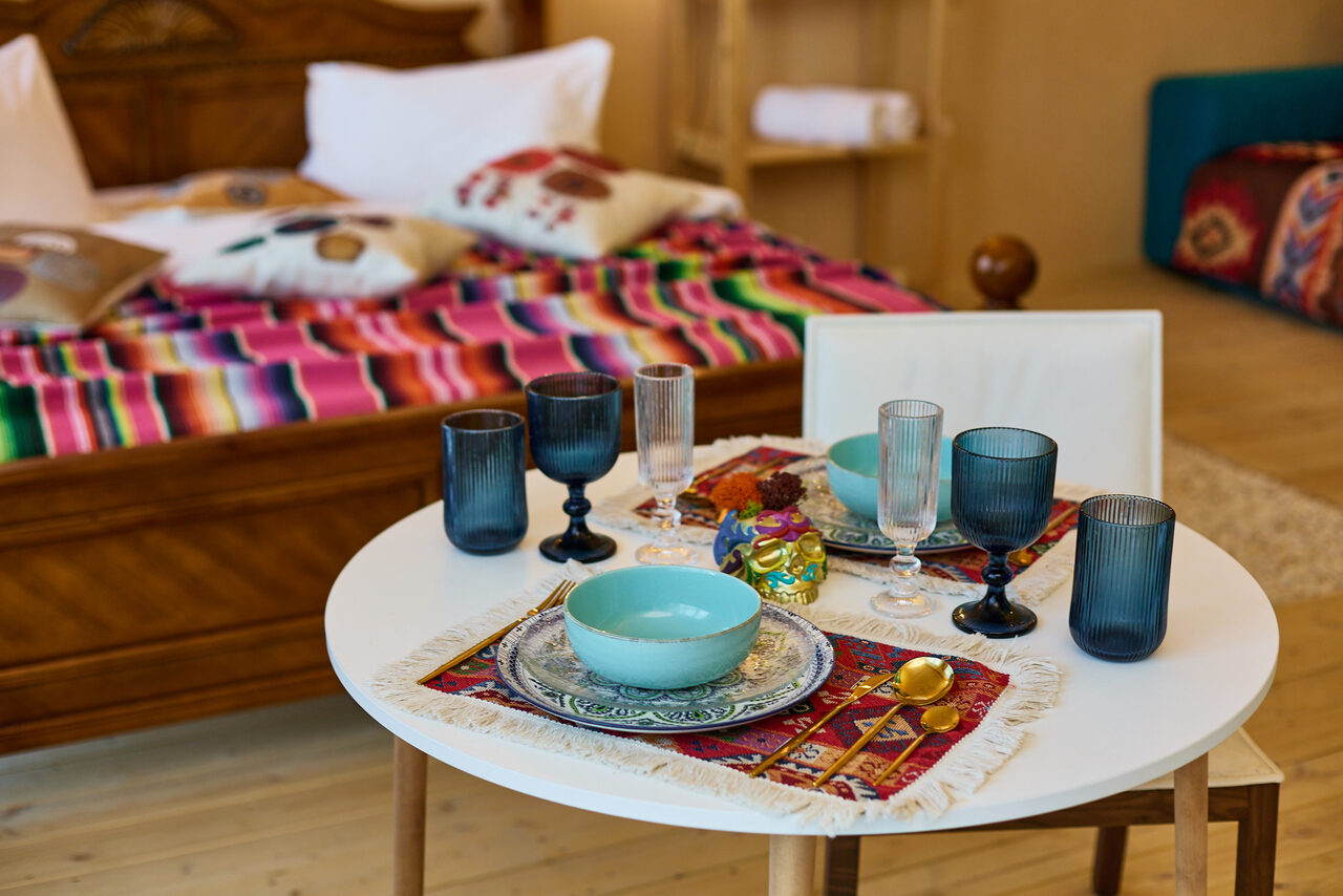 Арендовать ⁠Лофт в Мексиканском стиле для костюмированных вечеринок. Круглый обеденный стол с белой столешницей, сервирован цветной посудой, бокалами и столовыми приборами. На столе тканевая салфетка с узором. Внутреннее пространство с деревянным полом, кровать с ярким покрывалом и подушками в фоне.