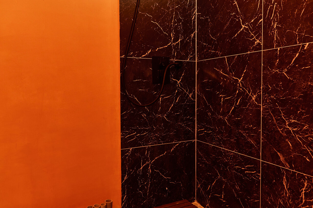 Арендовать ⁠Лофт в Мексиканском стиле для костюмированных вечеринок. Стена из оранжевой штукатурки и угловая плитка с черным мраморным узором. Потайное освещение создает теплый оттенок. Видны элементы душевой системы.