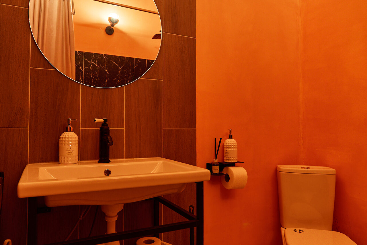 Арендовать ⁠Лофт в Мексиканском стиле для костюмированных вечеринок. Санузел с оранжевыми стенами и светло-коричневой плиткой. Установлены умывальник с тёмным смесителем, зеркало круглой формы, диспенсер, туалетная бумага, ёршик и унитаз. Освещение от настенного светильника.